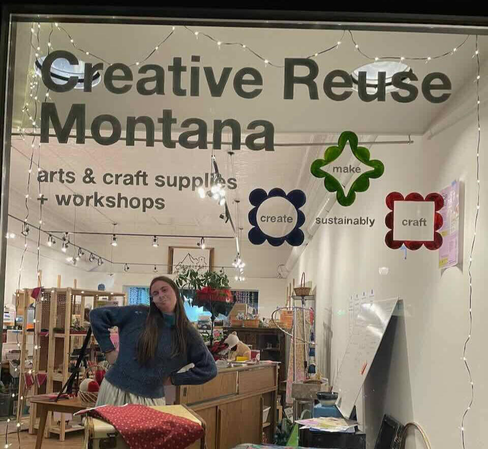 Creative Reuse Montana Image shows a store window with the words "Creative Reuse Montana" in bold text across the top and a view into a brightly lit store with art supplies on shelves and a friendly person with long brown hair and a blue sweater striking a pose